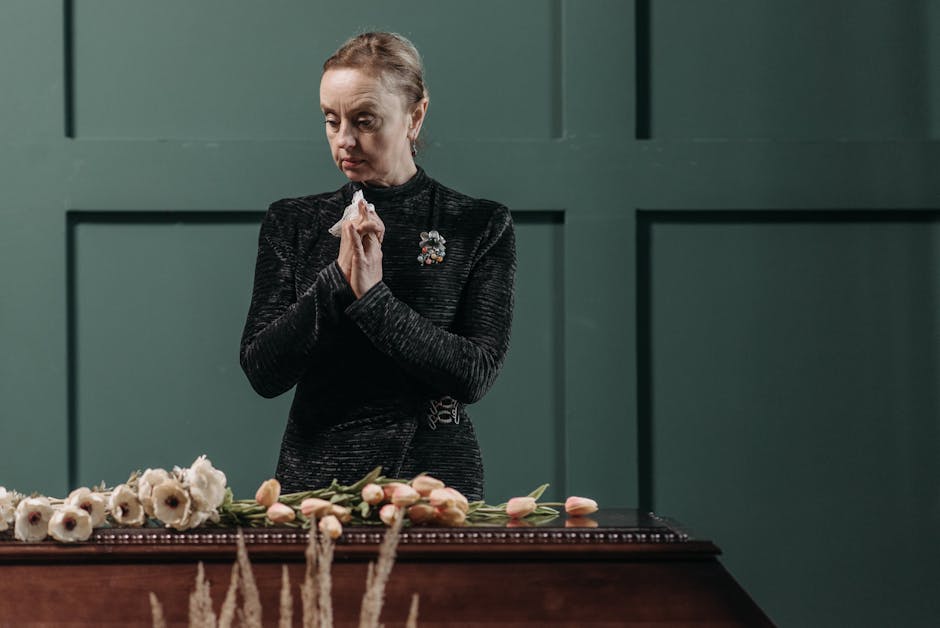 A woman dressed in a black textured long-sleeve top stands solemnly behind a wooden table, her hands clasped in a prayerful gesture. In front of her, there is an arrangement of fresh funeral flowers,