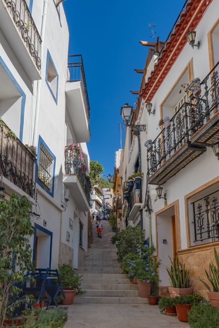 A narrow, ascending street in a picturesque village featuring whitewashed buildings with wrought iron balconies, some decorated with vibrant flower arrangements in shades of pink, purple, and red. The