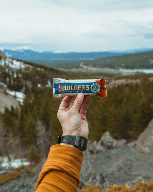 A close-up of a person's hand holding a protein bar labeled 'Builder's 20' with the background of a scenic outdoor landscape featuring rolling hills, scattered trees, rocky terrain, and distant mounta