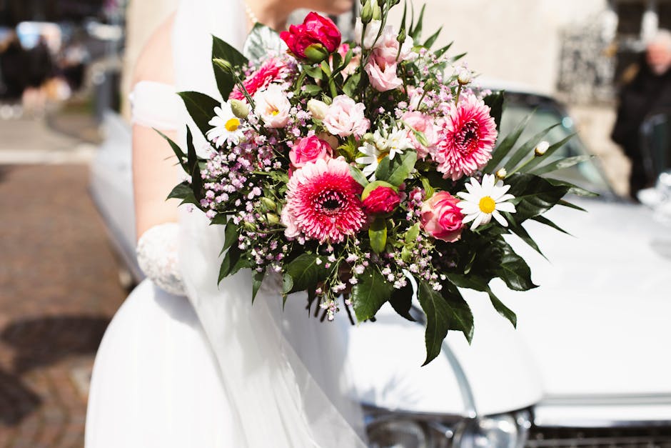 A bride holding a lush bouquet of fresh flowers, featuring large pink gerbera daisies, delicate pink eustoma, white daisies with yellow centers, and small clusters of pink baby's breath. The arrangeme